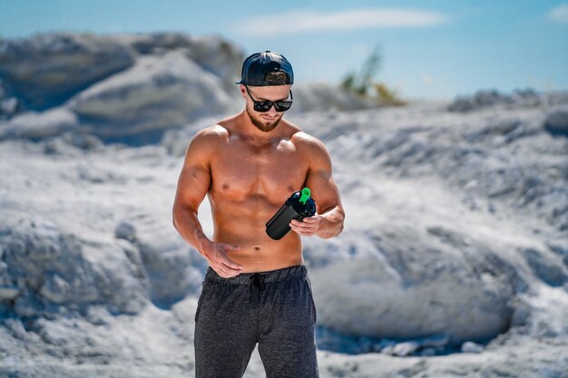 Portret van een knappe halfnacked bodybuilder man in glazen en een pet met een fles water.