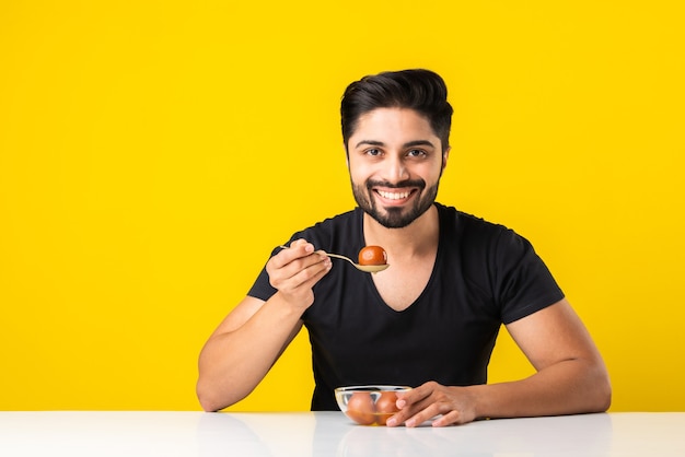 Portret van een knappe Indiase jongeman die zoete Gulab Jamun eet tegen een gele achtergrond