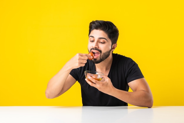 Portret van een knappe Indiase jongeman die zoete Gulab Jamun eet tegen een gele achtergrond