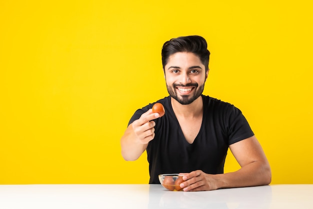 Portret van een knappe Indiase jongeman die zoete Gulab Jamun eet tegen een gele achtergrond