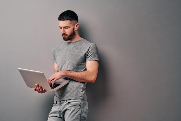 Portret van een knappe jonge man die tegen een grijze muur leunt en een laptop vasthoudt