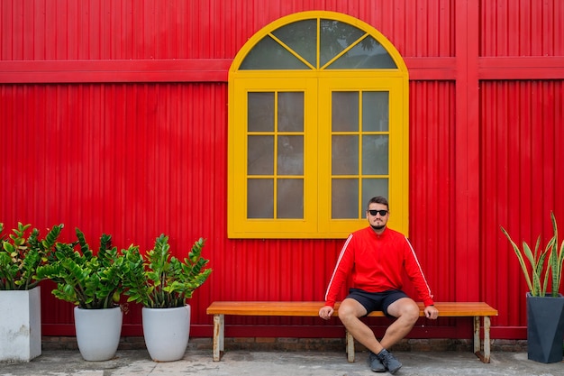 Foto portret van een knappe jongeman gekleed in een rood jasje en zwarte korte broek poseren tegen de achtergrond van een rood gebouw met een geel raam en ingemaakte bloemen