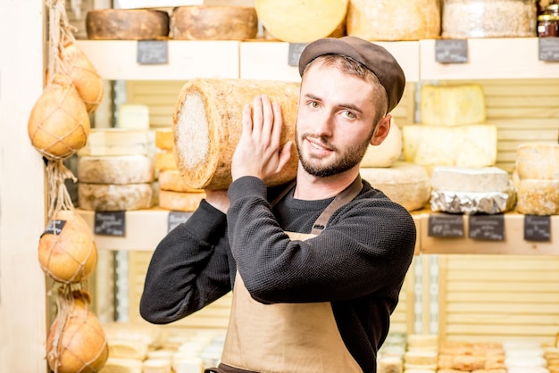Portret van een knappe kaasverkoper in uniform met een grote gekruide kaas voor de winkelvitrine vol met verschillende kazen