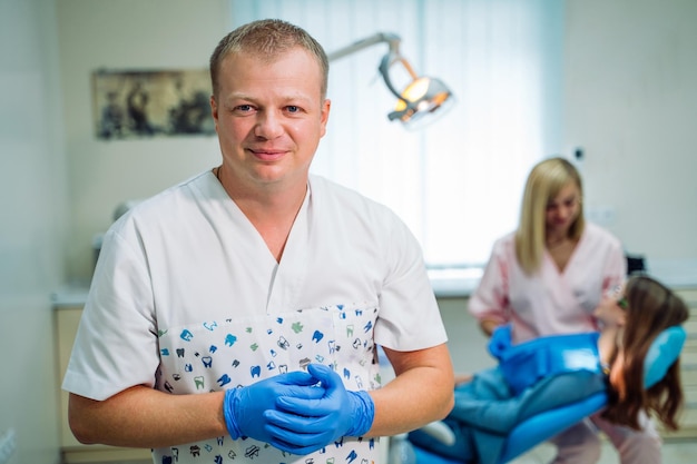 Portret van een knappe lachende tandarts met zijn verpleegster of helper die met de patiënt op de achtergrond werkt. Tandheelkunde concept