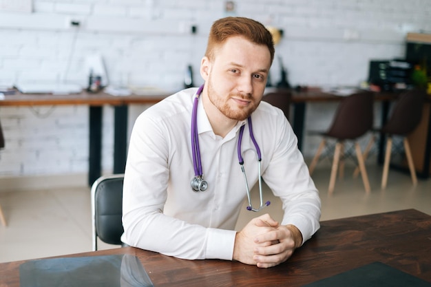 Portret van een knappe mannelijke arts die een wit uniform draagt met een stethoscoop die aan de balie in het ziekenhuiskantoor zit en naar de camera kijkt
