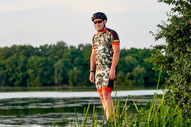 Portret van een knappe mannelijke fietser die in de buurt van de rivier staat tegen een prachtig landschap met een meer. Mens die weg kijkt.