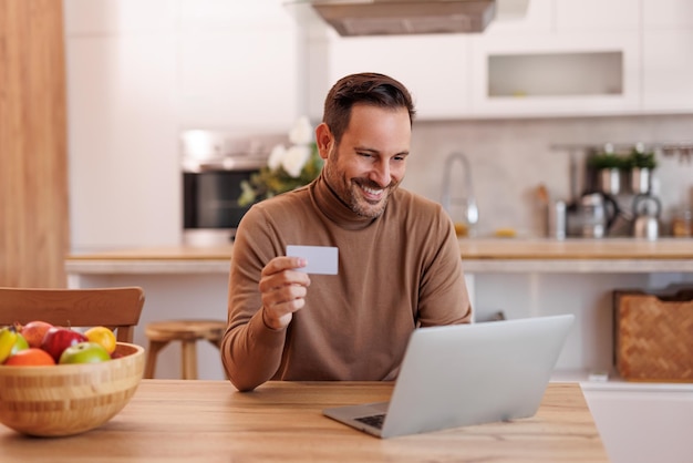 Portret van een knappe middelgrote man met een creditcard die online betaalt over een laptop eettafel