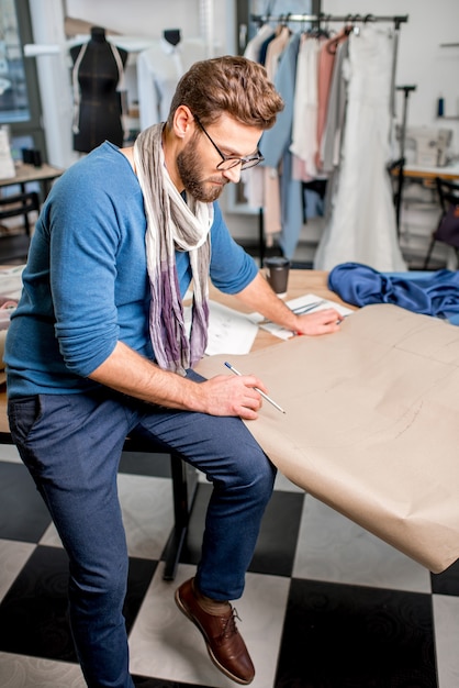 Portret van een knappe modeontwerper die met papieren schetsen zit in de studio vol kleermakersgereedschap en kleding