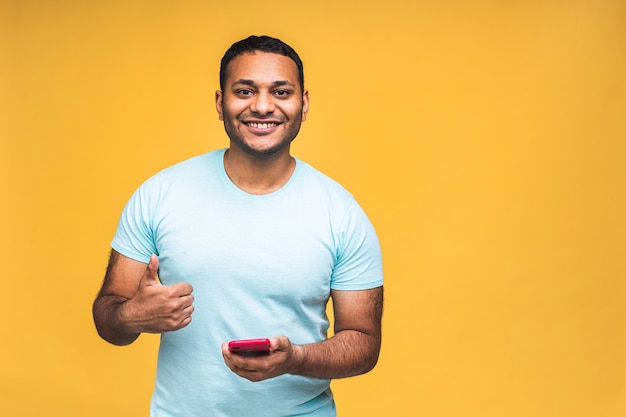 Portret van een knappe opgewonden vrolijke indiase Afro-Amerikaanse man die casual berichten naar zijn geliefde verzendt en ontvangt, geïsoleerd op gele achtergrond. Telefoon gebruiken. Duimen omhoog.