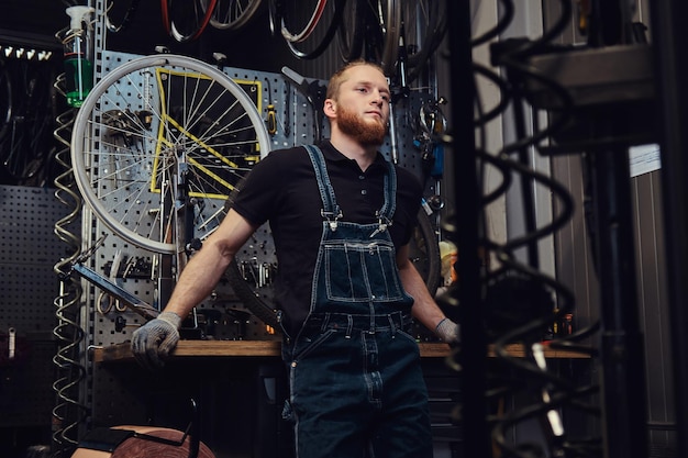 Portret van een knappe roodharige man met baard en kapsel in een spijkerbroek, staande in de buurt van een fietswiel in een werkplaats.