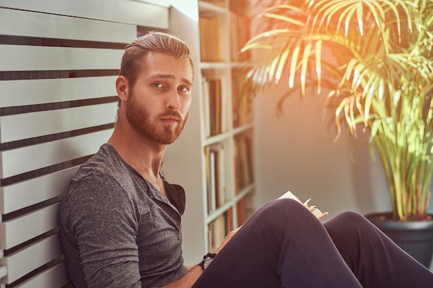 Portret van een knappe stijlvolle roodharige student in vrijetijdskleding, zittend op een vloer bij een huis en houdt een boek vast, kijkend naar een camera.