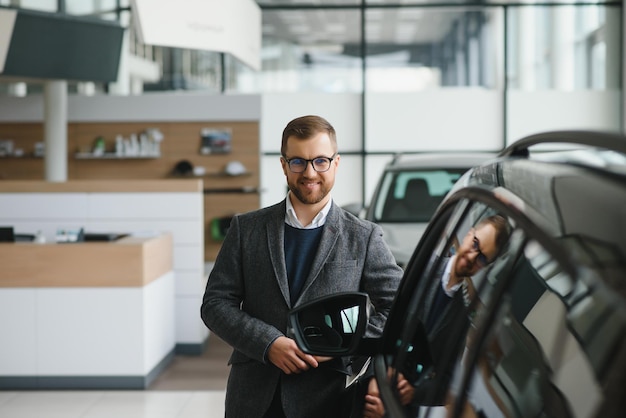 Portret van een knappe verkoper in het pak die bij de auto in de showroom staat