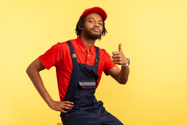 Portret van een knappe zelfverzekerde klusjesman met een blauw uniform, een rode vizierdop en een T-shirt die zich voordeed en duimen opsteekt, kijkend naar de camera. Indoor studio opname geïsoleerd op gele achtergrond.