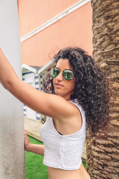 Portret van een krullende brunette surfer vrouw met witte top en zwarte bikini met een surfplank naast de palmboom