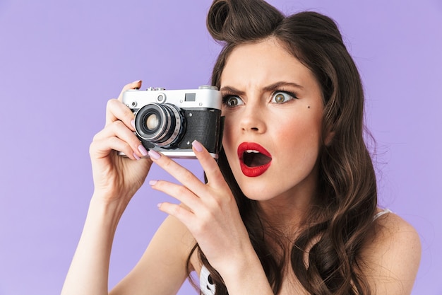 Portret van een krullende pin-up vrouw in vintage polka dot jurk die lacht terwijl ze een retro fotocamera vasthoudt die over een violette muur wordt geïsoleerd