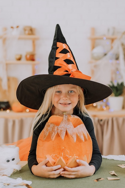 Portret van een lachend blond meisje in een enorme heksenhoed omhelst een groot halloween-concept met pompoenlantaarns