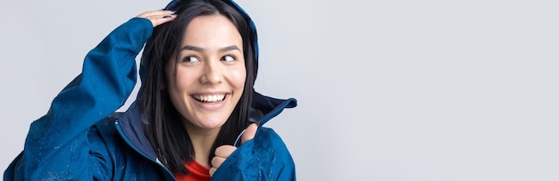 Portret van een lachend meisje gekleed in blauwe regenjas in druppels poseren met capuchon op grijze achtergrond in een studio