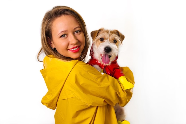 Portret van een lachend meisje in een gele regenjas met een hond Jack Russell Terrier in een rood jasje in haar armen. Geïsoleerd op witte achtergrond