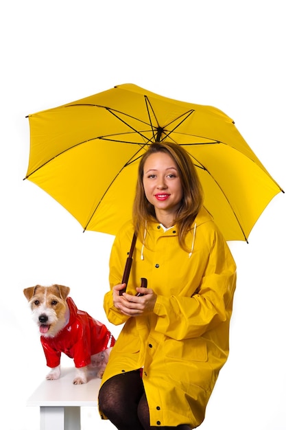 Portret van een lachend meisje in een gele regenjas met een hond Jack Russell Terrier in een rood jasje met een paraplu in zijn handen. Geïsoleerd op een witte achtergrond.