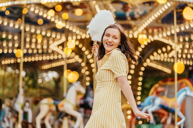 Portret van een lachend meisje met suikerspin voor de carrousel met nachtverlichting in het pretpark.