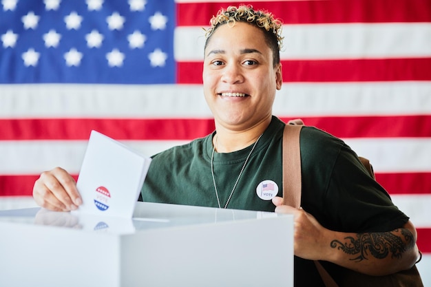 Portret van een lachende Afro-Amerikaanse vrouw die een stembiljet in de prullenbak stopt tegen een achtergrondkopie van de banner van de V.S