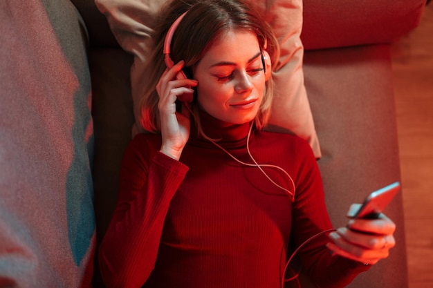 Portret van een lachende dame in een koptelefoon die op de bank ligt met een mobiele telefoon in de hand en thuis muziek luistert