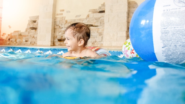 Portret van een lachende en lachende 3 jaar oude kleine jongen die zwemt met een opblaasbare kleurrijke ring en speelt met een strandbal in het zwembad