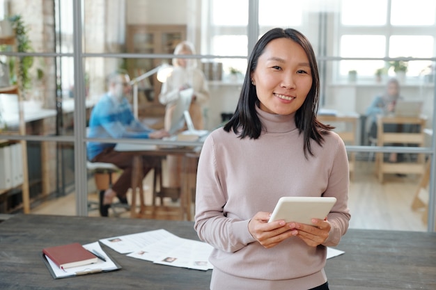 Portret van een lachende jonge Aziatische HR-manager met een tablet die tegen een houten tafel staat met cv's van kandidaten