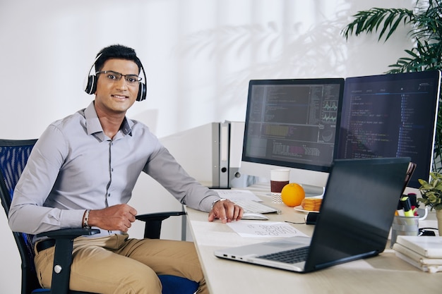 Portret van een lachende jonge Indiase softwareontwikkelaar in een bril die aan een bureau zit met computers en laptop