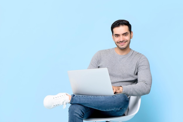 Portret van een lachende jonge knappe blanke man die naar de camera kijkt terwijl hij zit met een laptop in een geïsoleerde studio blauwe achtergrond