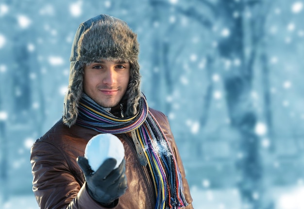 Portret van een lachende jonge man in een hoed met oorflap en een sneeuwbal in zijn hand