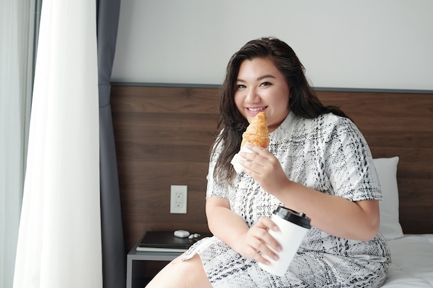 Portret van een lachende jonge vrouw die op bed zit en een croissant eet met koffie als ontbijt