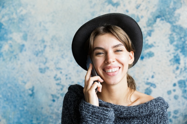 Portret van een lachende jonge vrouw gekleed in hoed en trui close-up praten op mobiele telefoon over blauwe muur