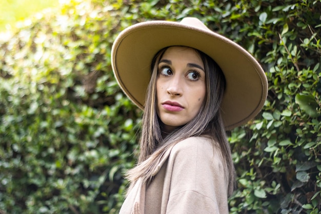 Portret van een lachende jonge vrouw in een hoed met groene bladeren op de achtergrond buitenshuis