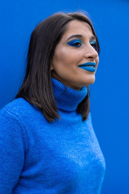 Portret van een lachende jonge vrouw met blauwe oogmake-up en lippenstift en blauwe trui op een blauwe achtergrond