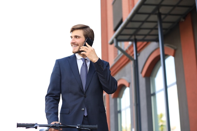 Portret van een lachende jonge zakenman gekleed in pak praten op mobiele staande buitenshuis.