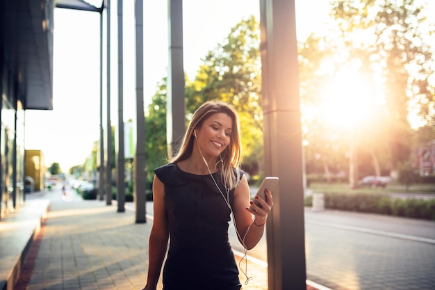 Portret van een lachende jonge zakenvrouw met koptelefoon die mobiele telefoon buitenshuis gebruikt.