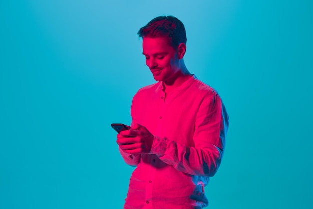 Portret van een lachende jongeman in een wit overhemd die berichten aan het typen is op een mobiele telefoon tegen blauw