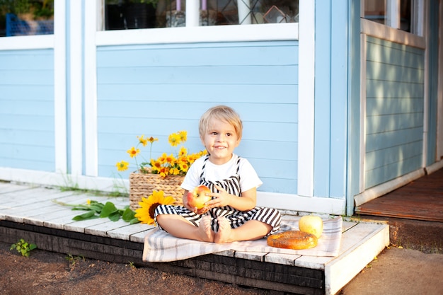 Portret van een lachende jongetje zittend op een houten veranda thuis op een herfstdag. Jeugd concept. Een kind speelt in de herfst in de tuin. Blij kind. Oogsten. Kleine boer. Charmante baby