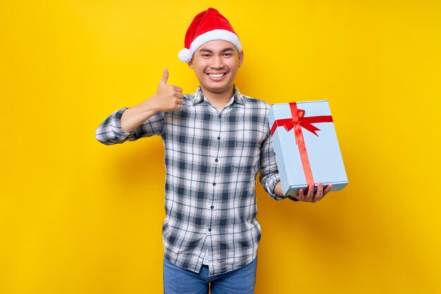 Portret van een lachende knappe jonge Aziatische man in een kerstman hoed met als cadeau Kerst gebaar geïsoleerd op gele studio achtergrond Merry Christmas Concept