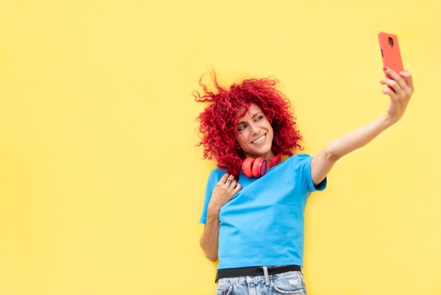 Portret van een lachende latijnse vrouw met rood afrohaar die een selfie neemt op een gele achtergrond met een blauwe t-shirt en rode koptelefoon om haar nek