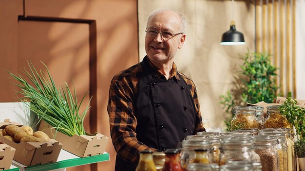 Portret van een lachende oude zero waste-supermarkteigenaar die afbreekbare glazen containers gebruikt voor zijn bulkartikelen om de impact op het klimaat te verminderen. Bejaarde man in de plaatselijke kruidenierswinkel die koopwaar regelt