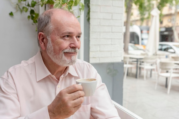 Portret van een lachende oudere man die uit het raam kijkt en geniet van een kopje koffie