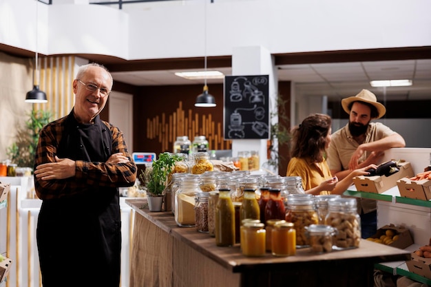 Portret van een lachende oudere verkoper in een zero waste-winkel naast biologisch afbreekbare, milieuvriendelijke voorraadpotten. Oudere man aan het werk in een milieuverantwoorde lokale buurtwinkel