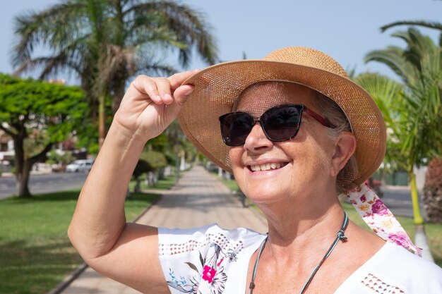 Portret van een lachende oudere vrouw met een strohoed en zonnebril in een openbaar park