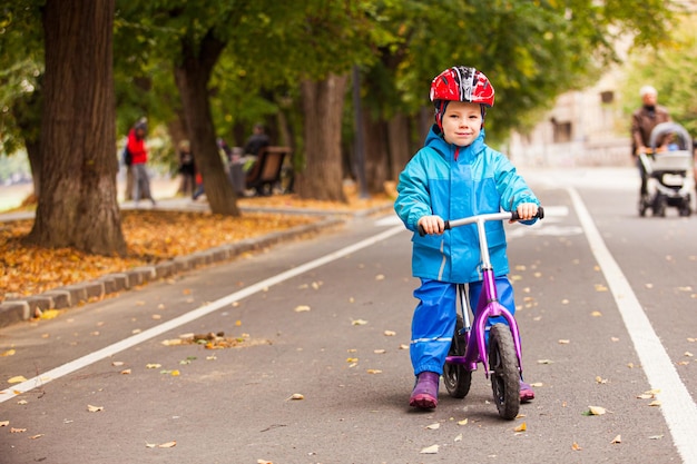 Portret van een lachende peuterjongen in een blauw herfst winddicht pak dat op zijn kleine fiets op straat zit te kijken naar de camera