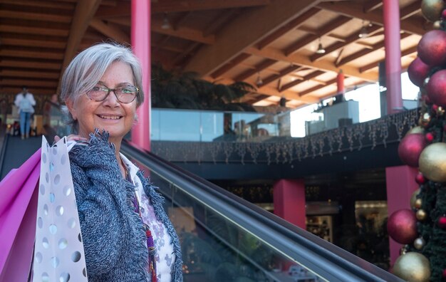 Portret van een lachende senior vrouw met wit haar die boodschappen doet voor kerstmis in een winkelcentrum, terwijl ze veel tassen vasthoudt met cadeaus voor familie en vrienden. consumentisme concept