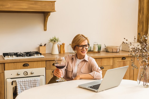 Portret van een lachende volwassen senior vrouw die een glas wijn vasthoudt terwijl ze een laptop op de keukentafel gebruikt. Freelance thuiswerken concept.