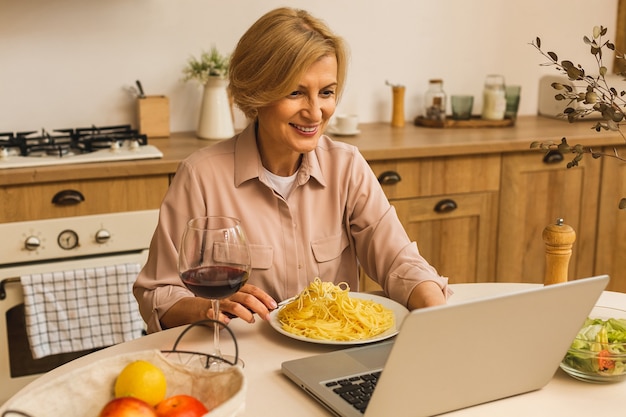 Portret van een lachende volwassen senior vrouw die een glas wijn vasthoudt terwijl ze een laptop op de keukentafel gebruikt. Freelance thuiswerken concept.