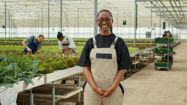 Portret van een lachende vrouw in een moderne kas met werknemers die voor slateelt en zaailingen van groene planten zorgen. afro-amerikaanse landarbeider poseren gelukkig in biologische groenten boerderij.